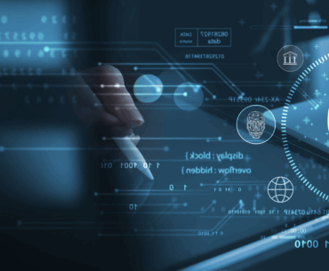 Closeup of a pair of hands. One is typing on a laptop and the other is drawing on a tablet with a stylus. There are various holograms in the foreground, most prominently a shield with a lock in the middle.