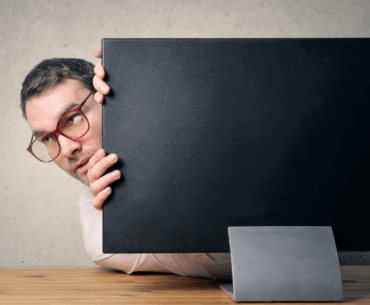 A man wearing glasses is poking his head around a computer monitor as if he's hiding. This is a metaphor for how to hide my IP address.