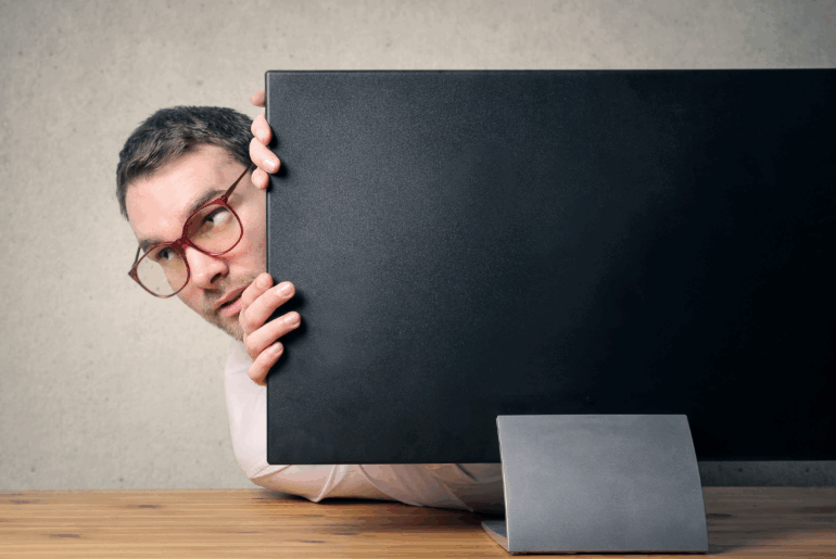 A man wearing glasses is poking his head around a computer monitor as if he's hiding. This is a metaphor for how to hide my IP address.