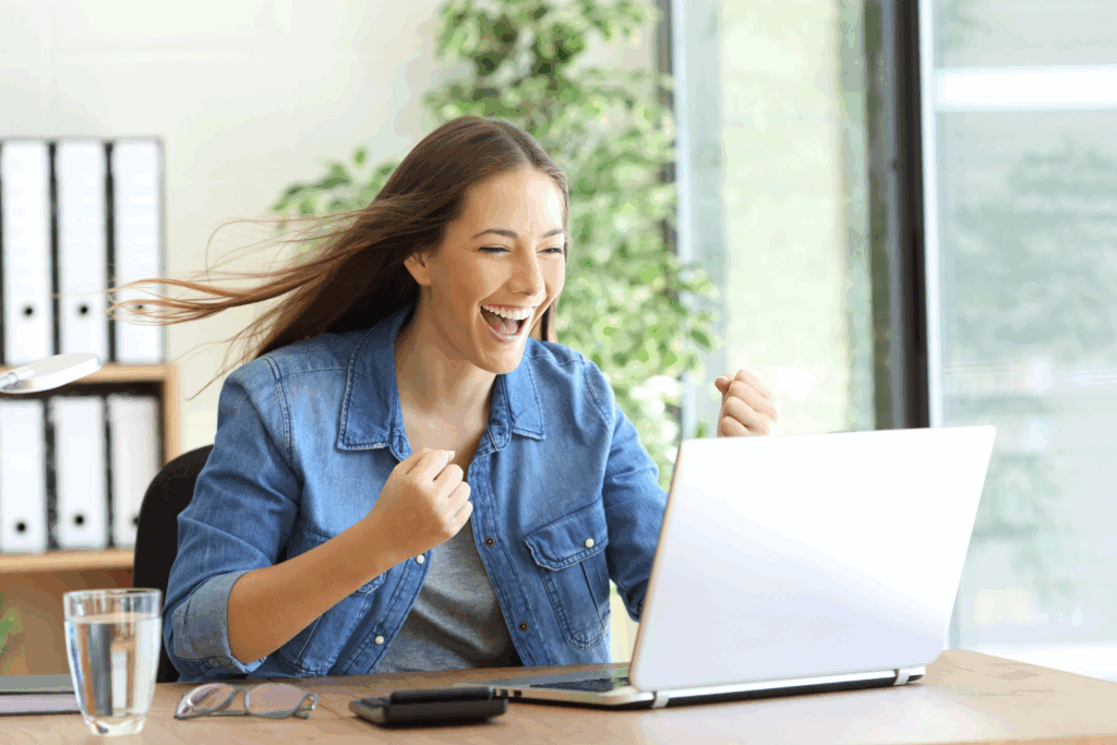 A woman wearing a blue button-down shirt with her hair flying out behind her is celebrating in front of her laptop because of how fast her Internet connection is.