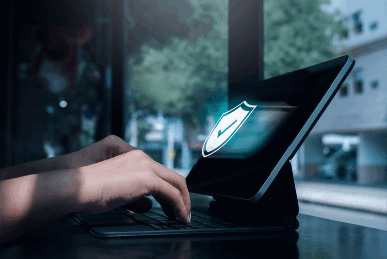 Closeup of a pair of hands typing on a laptop. There is a shield with a checkmark on it that seems to be coming out of the screen .