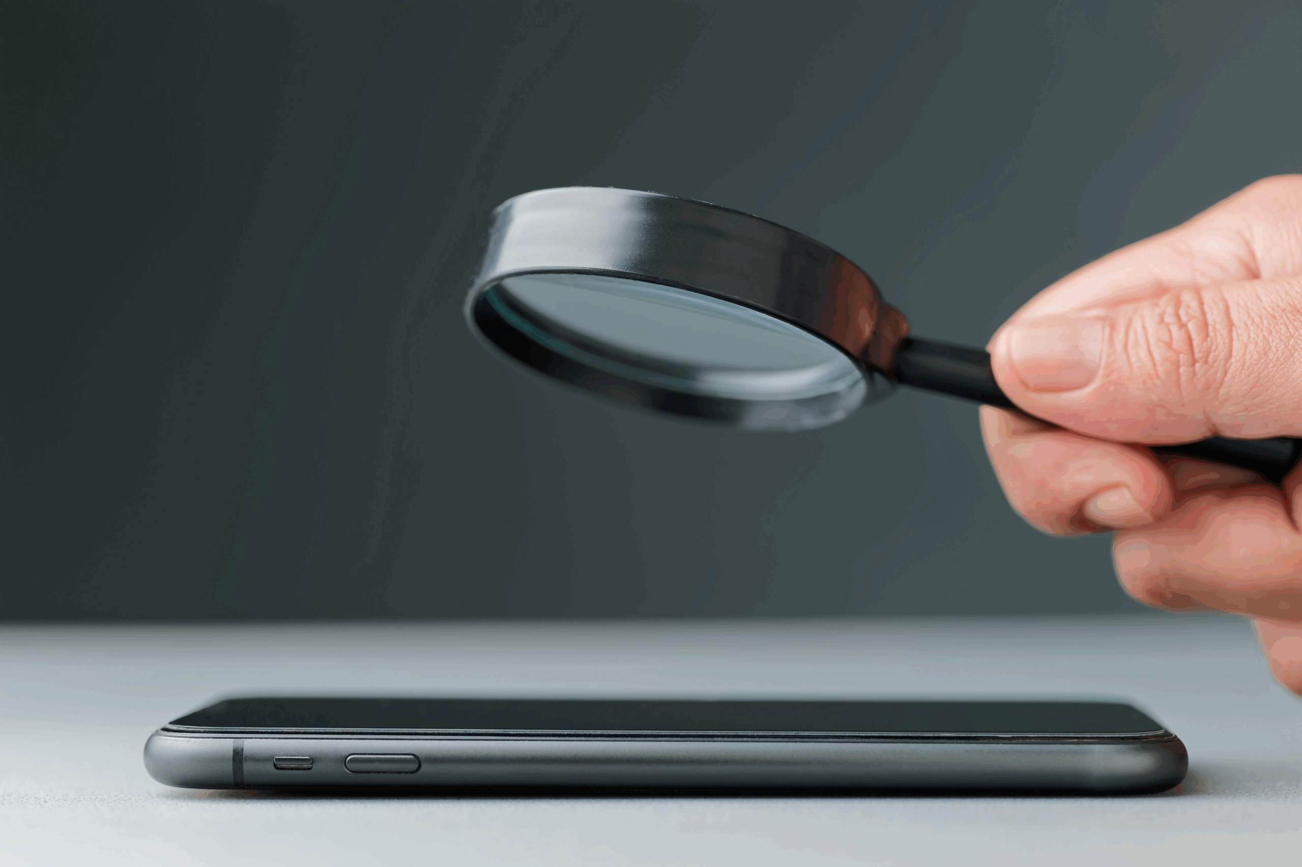 Closeup of a hand holding a magnifying glass over a smartphone, looking for security breaches that can be solved with PrivadoVPN.