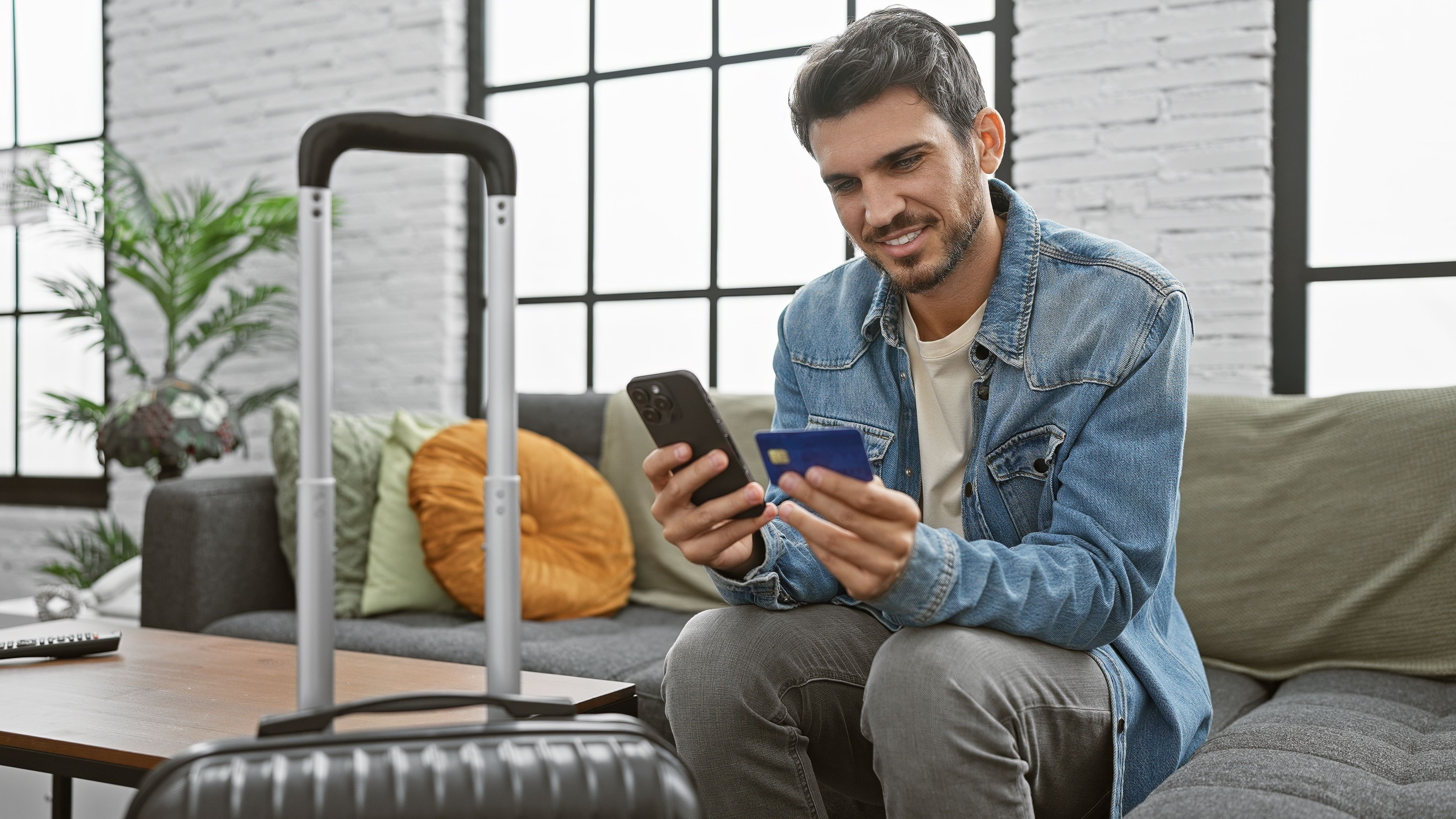 A man in a jean jacket is sitting on a couch. He is purchasing PrivadoVPN Premium on a burner phone. His suitcase is in the foreground, so he's about to travel and wants his data to stay secure.