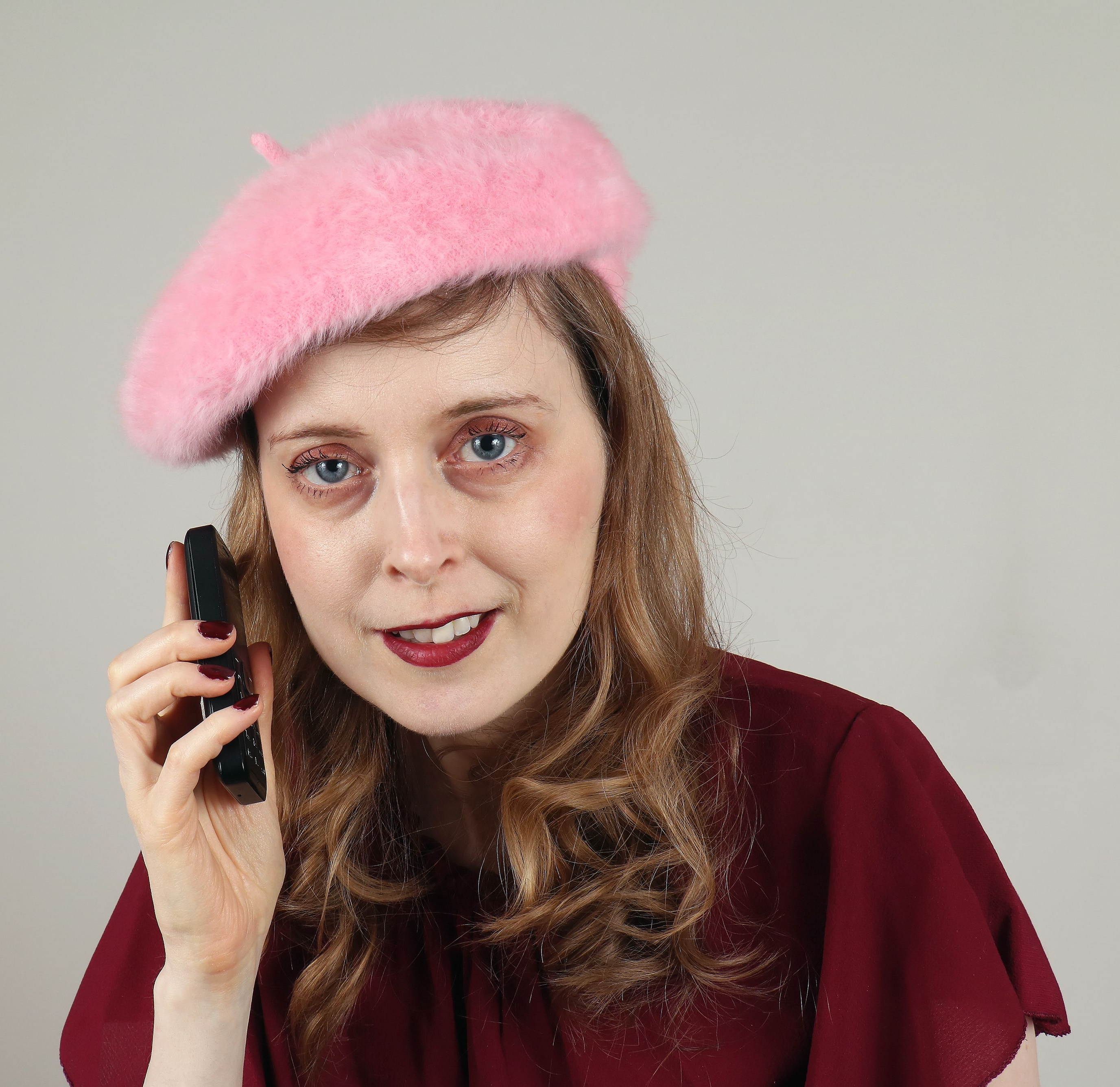 A woman who looks like a young Carol Kane is wearing a pink, fuzzy beret and talking on a burner phone.